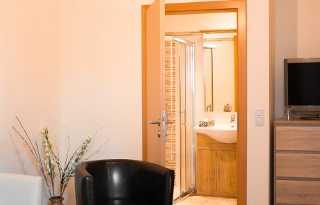 IMG_2326 Wohnzimmer mit zwei Sesseln, einem dekorativen Beistelltisch, einer Kommode, einem Fernseher und geöffneter Tür zum Badezimmer
